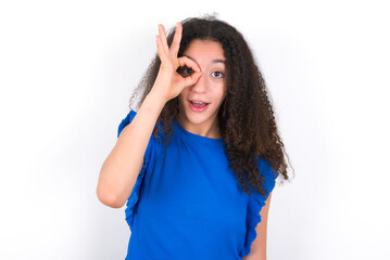 Fototapeta premium Teenager girl with afro hairstyle wearing blue T-shirt over white wall doing ok gesture shocked with surprised face, eye looking through fingers. Unbelieving expression.