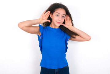 Obraz na płótnie Canvas Teenager girl with afro hairstyle wearing blue T-shirt over white wall Trying to hear both hands on ear gesture, curious for gossip. Hearing problem, deaf