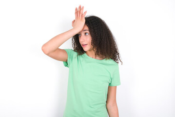 Teenager girl with afro hairstyle wearing green T-shirt over white wall surprised with hand on head for mistake, remember error. Forgot, bad memory concept.