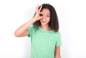 Teenager girl with afro hairstyle wearing green T-shirt over white wall doing ok gesture with hand smiling, eye looking through fingers with happy face.