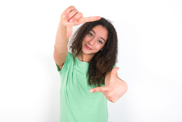 Teenager girl with afro hairstyle wearing green T-shirt over white wall making finger frame with hands. Creativity and photography concept.