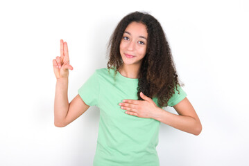 Teenager girl with afro hairstyle wearing green T-shirt over white wall smiling swearing with hand...