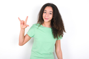 Teenager girl with afro hairstyle wearing green T-shirt over white wall doing a rock gesture and smiling to the camera. Ready to go to her favorite band concert.