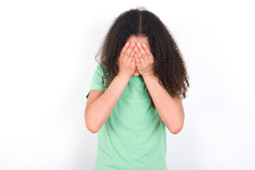 Teenager girl with afro hairstyle wearing green T-shirt over white wall covering her face with her hands, being devastated and crying. Sad concept