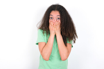Vivacious Teenager girl with afro hairstyle wearing green T-shirt over white wall, giggles joyfully, covers mouth, has natural laughter, hears positive story or funny anecdote