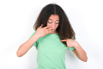 Shocked Teenager girl with afro hairstyle wearing green T-shirt over white wall look surprisedly...