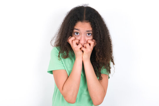 Fearful Teenager girl with afro hairstyle wearing green T-shirt over white wall keeps hands near mouth, feels frightened and scared,  has a phobia,  Shock and frighted concept.