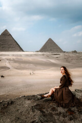 Young redhead tourist girl in brown dress sitting on a stone cliff in Egypt, Cairo - Giza. Pyramids on backround. Copy space