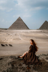 Young redhead tourist girl in brown dress sitting on a stone cliff in Egypt, Cairo - Giza. Pyramids on backround. Copy space