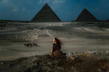 Young redhead tourist girl in brown dress sitting on a stone cliff in Egypt, Cairo - Giza. Pyramids on backround and dramatic light. Copy space - obrazy, fototapety, plakaty