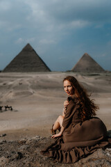 Young redhead tourist girl in brown dress sitting on a stone cliff in Egypt, Cairo - Giza. Pyramids on backround. Copy space