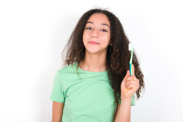 Teenager girl with afro hairstyle wearing green T-shirt over white wall holding a toothbrush and smiling. Dental healthcare concept.