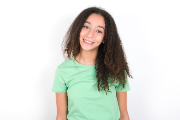 Fototapeta premium Teenager girl with afro hairstyle wearing green T-shirt over white wall with nice beaming smile pleased expression. Positive emotions concept