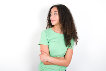 Pleased Teenager girl with afro hairstyle wearing green T-shirt over white wall keeps hands crossed over chest looks happily aside