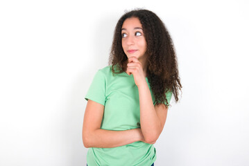 Thoughtful Teenager girl with afro hairstyle wearing green T-shirt over white wall holds chin and looks away pensively makes up great plan