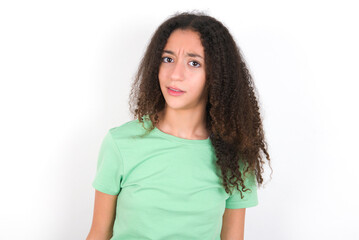 Portrait of dissatisfied Teenager girl with afro hairstyle wearing green T-shirt over white wall smirks face, purses lips and looks with annoyance at camera, discontent hearing something unpleasant