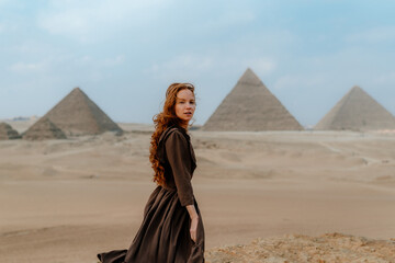 Young redhead tourist girl wearing a brown dress standing on the sand in Egypt, Cairo - Giza. Pyramids on backround. Copy space - obrazy, fototapety, plakaty