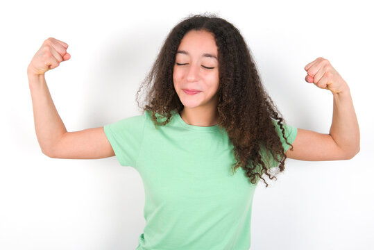 Strong powerful Teenager girl with afro hairstyle wearing green T-shirt over white wall toothy smile, raises arms and shows biceps. Look at my muscles!