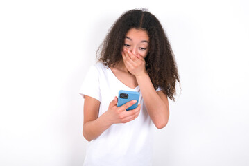 Teenager girl with afro hairstyle wearing white T-shirt over white wall  being deeply surprised,...