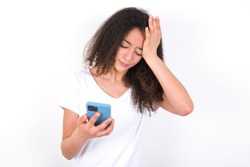 Upset depressed Teenager girl with afro hairstyle wearing white T-shirt over white wall  makes face palm as forgot about something important holds mobile phone expresses sorrow and regret blames