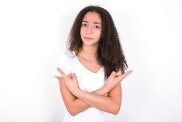 Serious Teenager girl with afro hairstyle wearing white T-shirt over white wall  crosses hands and points at different sides hesitates between two items. Hard decision concept