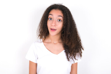 Shocked Teenager girl with afro hairstyle wearing white T-shirt over white wall  stares bugged eyes keeps mouth opened has surprised expression. Omg concept