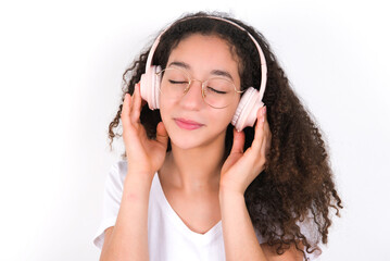 Fototapeta premium Pleased Teenager girl with afro hairstyle wearing white T-shirt over white wall enjoys listening pleasant melody keeps hands on stereo headphones closes eyes. Spending free time with music