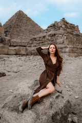 Young redhead tourist girl in brown dress sitting on a stone in Egypt, Cairo - Giza. Pyramids on backround. Copy space