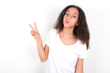 Teenager girl with afro hairstyle wearing white T-shirt over white wall  makes peace gesture keeps lips folded shows v sign. Body language concept