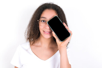 Teenager girl with afro hairstyle wearing white T-shirt over white wall  holding modern smartphone covering one eye while smiling