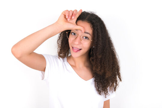 Funny Teenager girl with afro hairstyle wearing white T-shirt over white wall  makes loser gesture mocking at someone sticks out tongue making grimace face.
