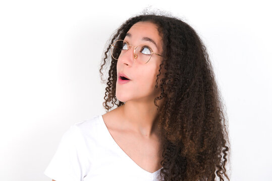 Shocked Teenager girl with afro hairstyle wearing white T-shirt over white wall  look empty space with open mouth screaming: Oh My God! I can't believe this.