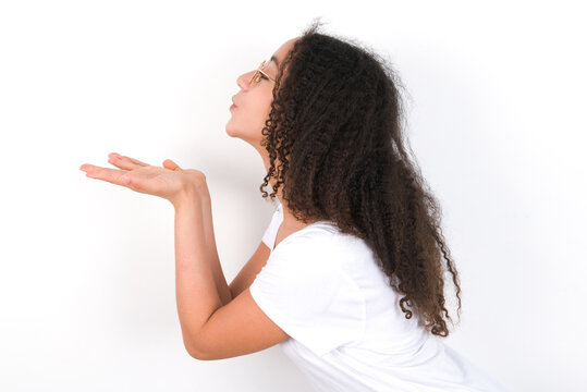 Profile side view view portrait of attractive Teenager girl with afro hairstyle wearing white T-shirt over white wall  sending air kiss