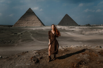 Young redhead tourist girl wearing a brown dress standing on the sand in Egypt, Cairo - Giza. Pyramids on backround. Copy space