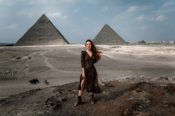 Young redhead tourist girl wearing a brown dress standing on the sand in Egypt, Cairo - Giza. Pyramids on backround. Copy space