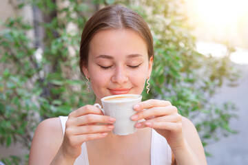 beautiful attractive happy positive smiling girl, young woman sitting outdoors on terrace in cafe on table smelling with closed eyes enjoying drinking beverage tea aroma coffee. Good summer morning