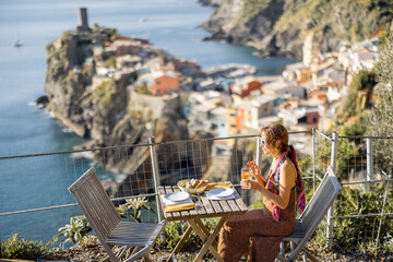 Woman sitting at outdoor restaurant with beautiful landscape of coastline in Vernazza, traveling in famous Cinque Terre region in northwestern Italy. Idea of mediterranean food and lifestyle