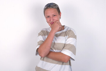 Thoughtful smiling young woman with short hair wearing striped t-shirt over white background keeps hand under chin, looks directly at camera, listens something with interest. Youth concept.