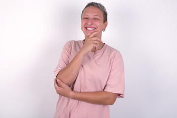 young woman with short hair wearing pink t-shirt over white background laughs happily keeps hand on chin expresses positive emotions smiles broadly has carefree expression