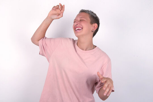 Photo of upbeat young woman with short hair wearing pink t-shirt over white background has fun and dances carefree wear being in perfect mood makes movements. Spends free time on disco party