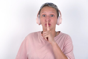Fototapeta na wymiar young woman with short hair wearing pink t-shirt over white background making hush gesture with finger on her lips wearing wireless headphones. Be quiet.