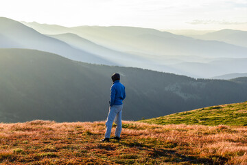 Naklejka premium A man stands in the mountains in a sunlit glade at sunset, back view