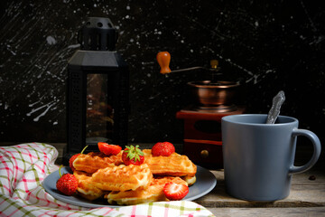 Waffles with strawberries and honey, sweet pastries plate and coffee mug, against background coffee grinder and kerosene lamp.