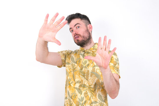 Portrait of smiling Young caucasian man wearing Hawaiian t-shirt over white background  looking at camera and gesturing finger frame. Creativity and photography concept.