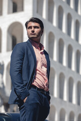 Businessman in shirt and jacket outdoors. Hands in the pocket. Behind him a building and the blue sky. Look into the distance.