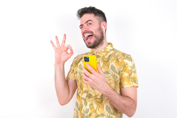 Happy young handsome bearded man wearing flowered shirt over white wall sending a message on his smartphone or taking a selfie  and making ok sign with his hand.