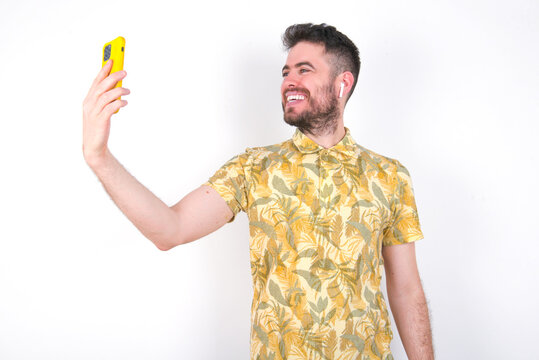 Portrait of a young handsome bearded man wearing flowered shirt over white wall  taking a selfie to send it to friends and followers or post it on his social media.