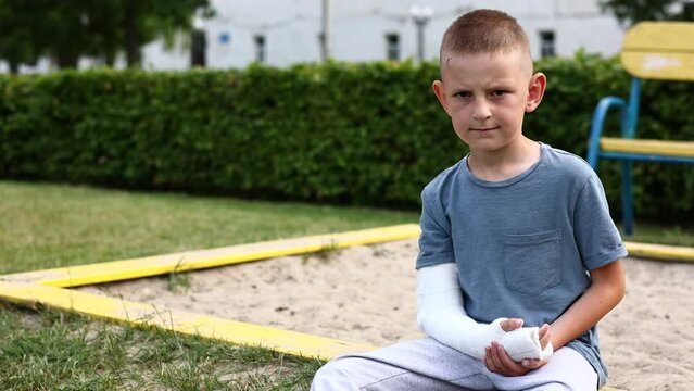 child caucasian with broken limb outdoors sits near the playground and looking on the plaster bandage on his arm. the worst summer vacation. concept of health accident, and medical.