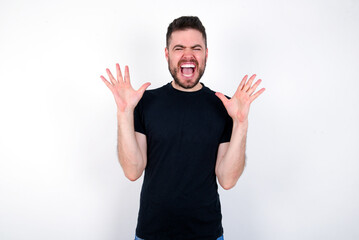 Fototapeta premium Crazy outraged young caucasian bearded man wearing black t-shirt standing over white wall screams loudly and gestures angrily yells furiously. Negative human emotions feelings concept