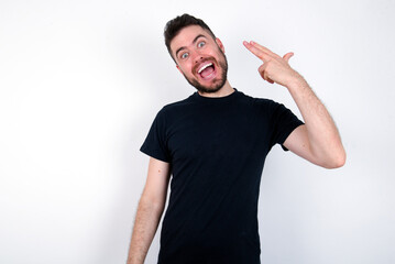 young caucasian bearded man wearing black t-shirt standing over white wall foolishness around shoots in temple with fingers makes suicide gesture. Funny model makes finger gun pistol